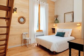 A bedroom with a wooden floor, a bed with white bedding, a chair, a window with white curtains, and a wall-mounted light fixture. - Eco-friendly gite in a wine estate in Frontignan (Frontignan)