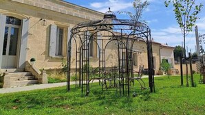 A garden with a metal gazebo, a stone building with a clock, and a clear blue sky.
