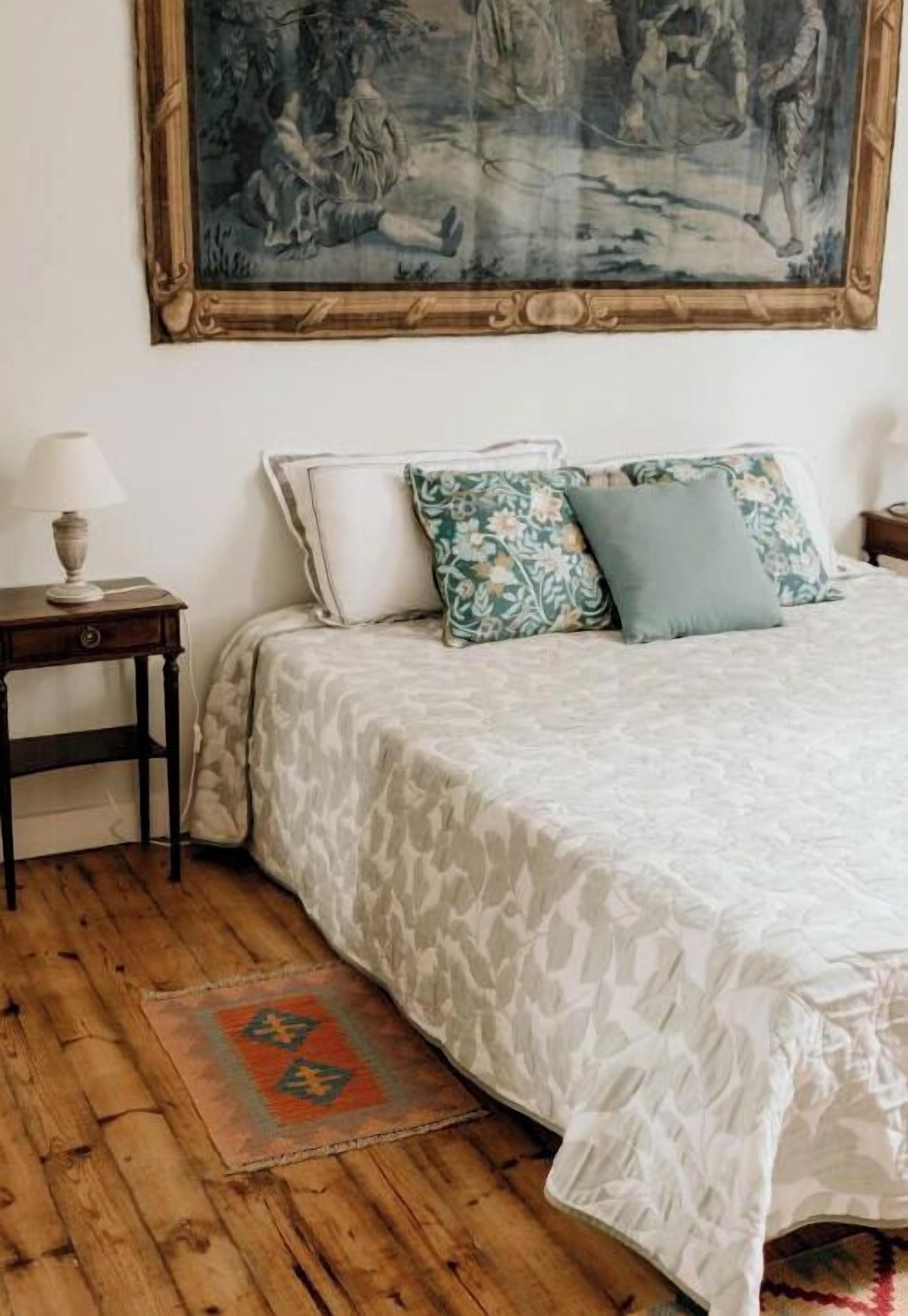 A bedroom with a bed, a nightstand, a lamp, a painting on the wall, and a patterned rug.