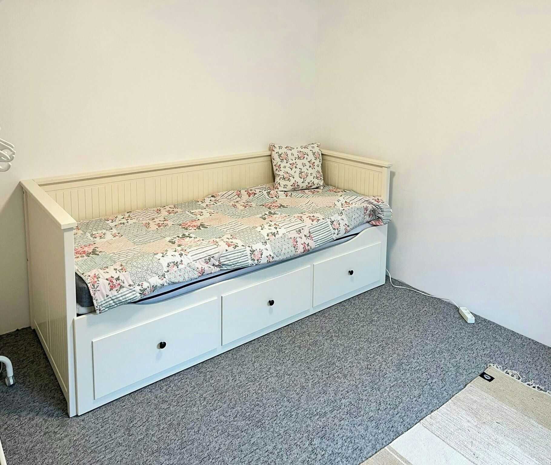 Une petite chambre avec un lit simple, une couvre-lit à motifs floraux, une tête de lit blanche et un sol recouvert de moquette.