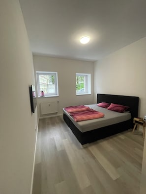 A bedroom with a bed, two windows, a radiator, and a small bedside table. - StayInn Suites | 100qm für 12 Gäste | Groß&Modern (Simbach am Inn)