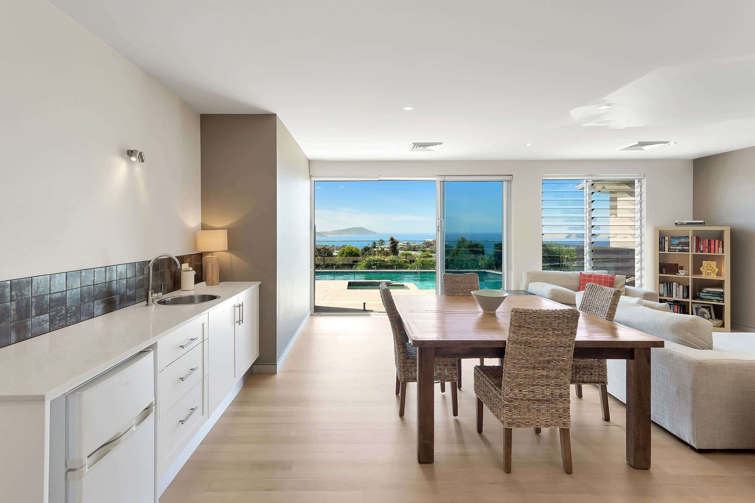 A modern kitchen with a large window, a dining area with a table and chairs, and a living room with a sofa and a bookshelf.
