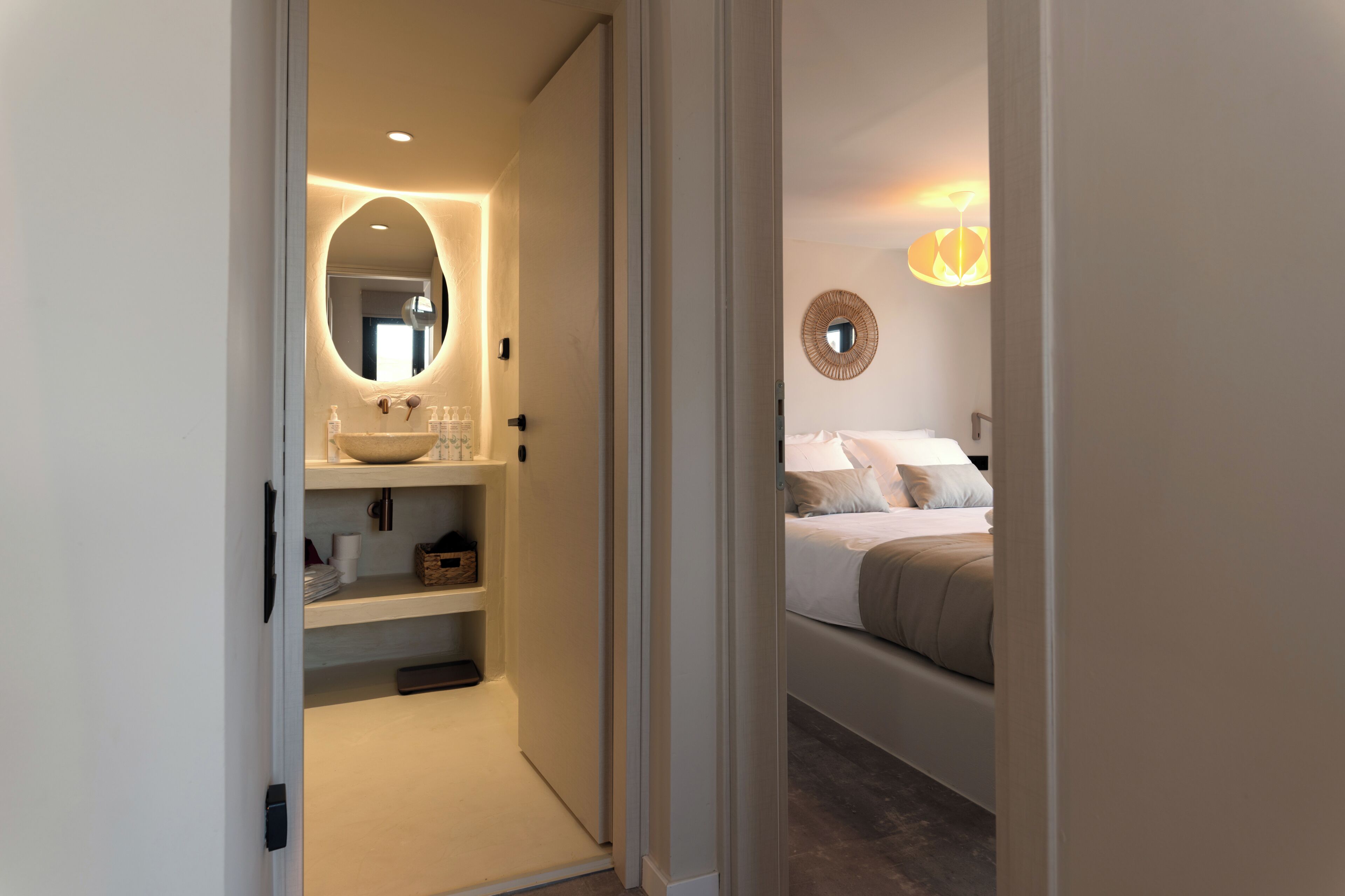 A hotel room with a bathroom featuring a large oval mirror, a sink with a vessel basin, and shelves with toiletries. The room has a bed with a headboard, pillows, and a bedside table.