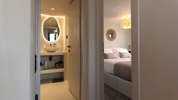 A hotel room with a bathroom featuring a large oval mirror, a sink with a vessel basin, and shelves with toiletries. The room has a bed with a headboard, pillows, and a bedside table.