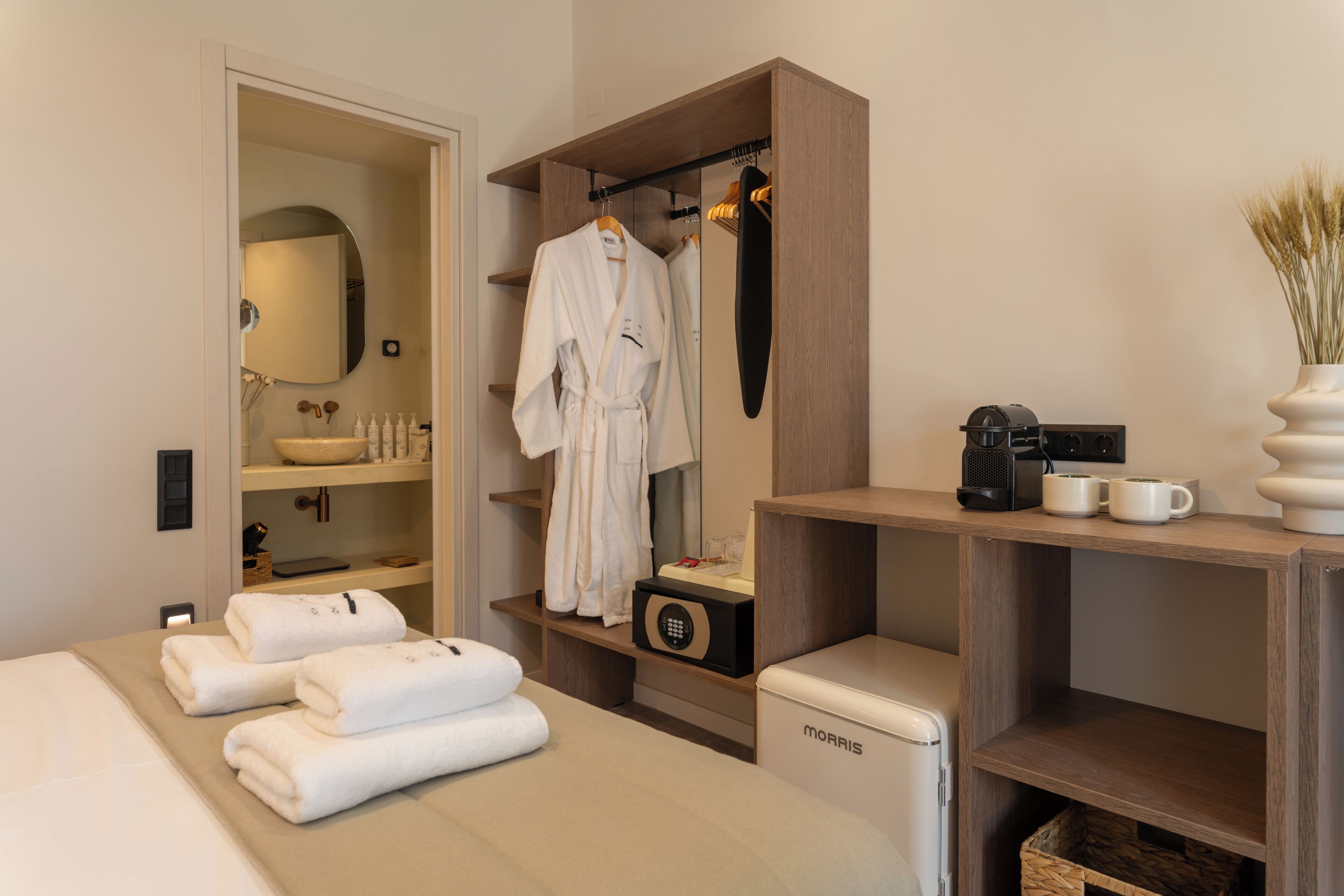 A modern hotel room with a bed, a bathroom with a sink and mirror, a robe hanging on a rack, a coffee maker, and a small refrigerator.
