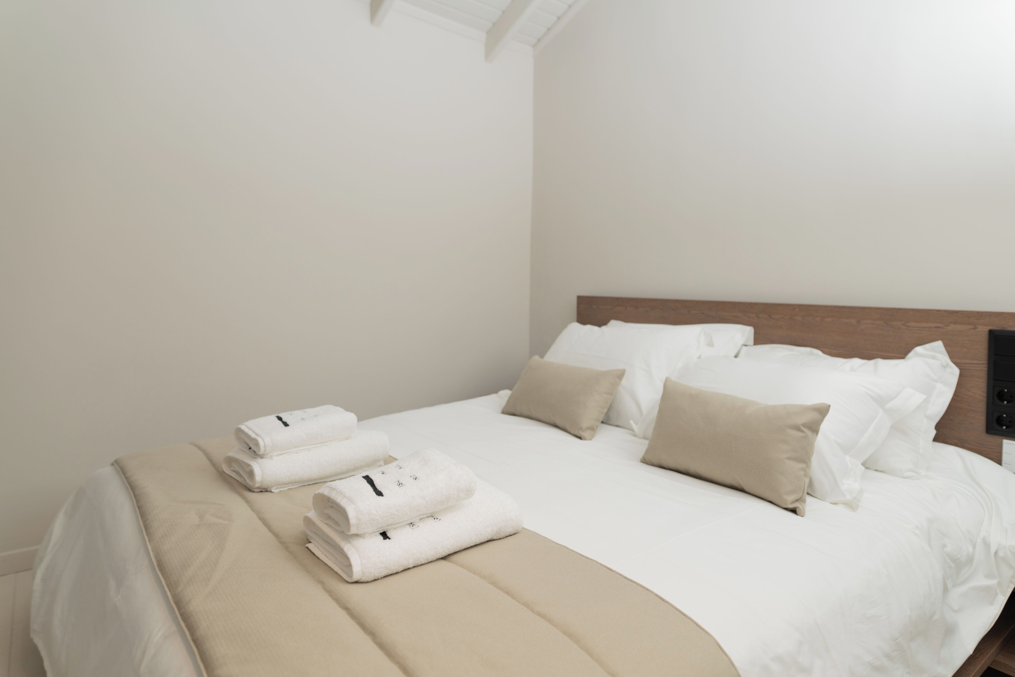 A neatly made bed with white linens and beige pillows, a wooden headboard, and a folded towel on the side.