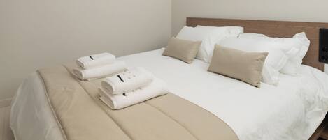 A neatly made bed with white linens and beige pillows, a wooden headboard, and a folded towel on the side.