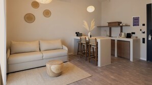 A modern living area with a sofa, a round wooden stool, a kitchen island with bar stools, and a pendant light. - Ichos Apartments (Volissos)