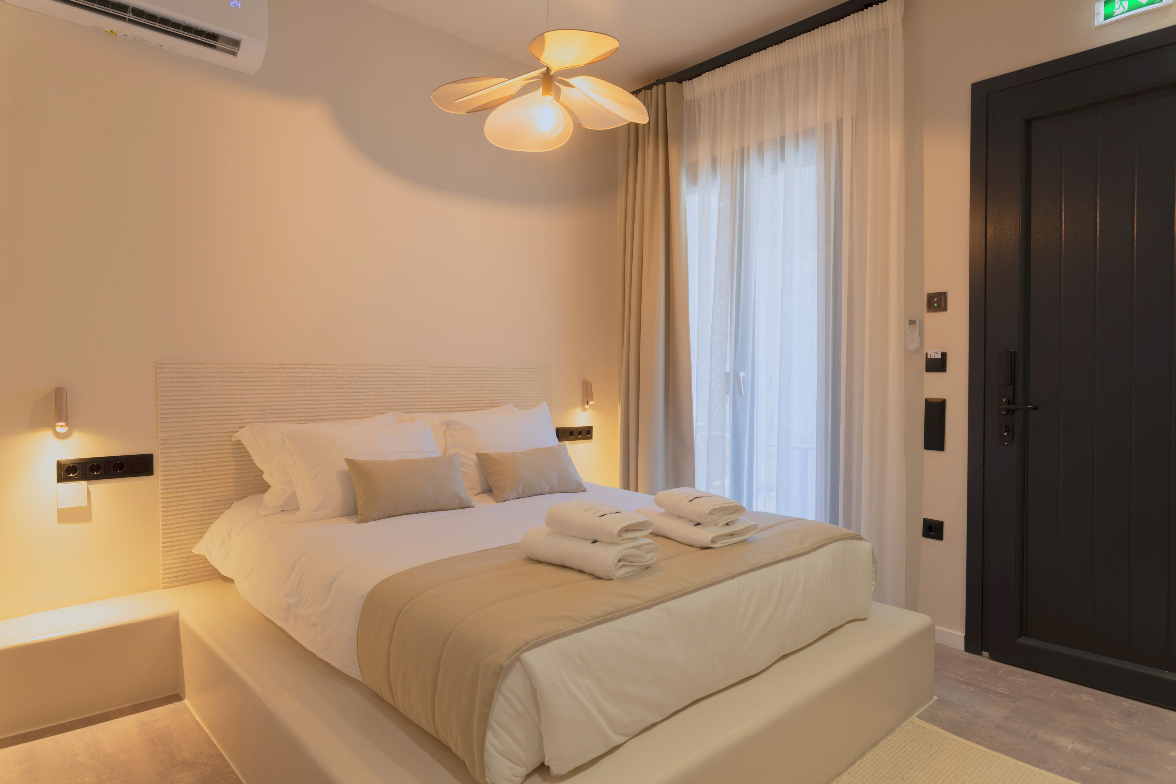 A hotel room with a bed, bedside lamps, a wall-mounted air conditioner, a door, and a window with curtains.