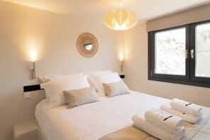 A hotel room with a large bed, two bedside lamps, a round mirror, and a window with a view. - Ichos Apartments (Volissos)