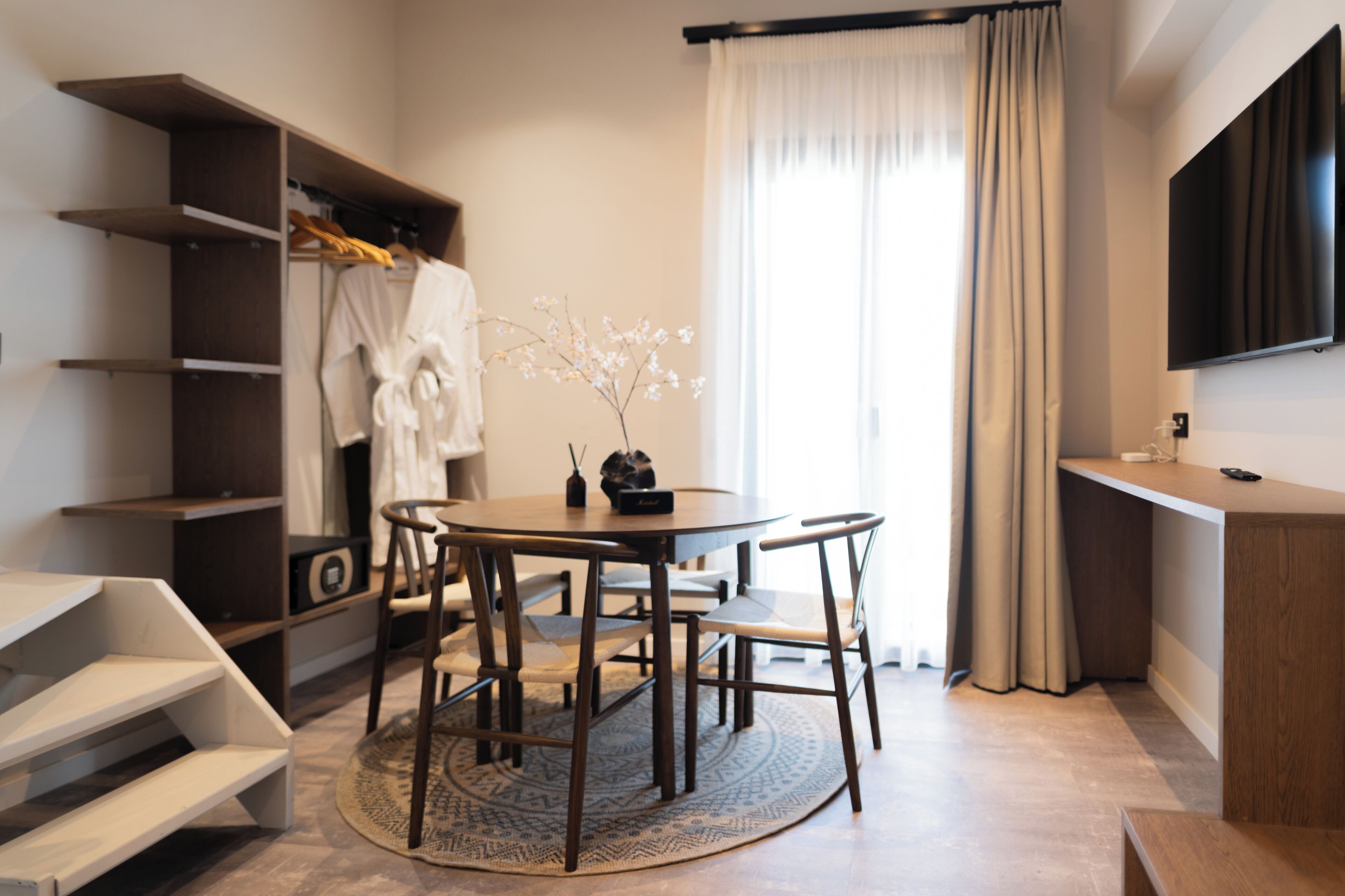 A modern hotel room with a round dining table, two chairs, a small side table, a TV mounted on the wall, a wardrobe with hanging clothes, and a large window with curtains.
