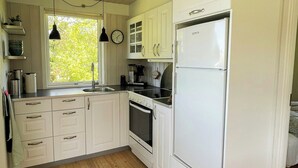 Une cuisine avec des meubles blancs, un réfrigérateur, un évier, une cuisinière et une fenêtre donnant sur des arbres.