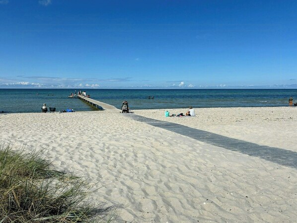 Ein Sandstrand mit einem Steg, der ins Wasser ragt, Menschen sitzen und gehen am Ufer entlang, und ein klarer, blauer Himmel.