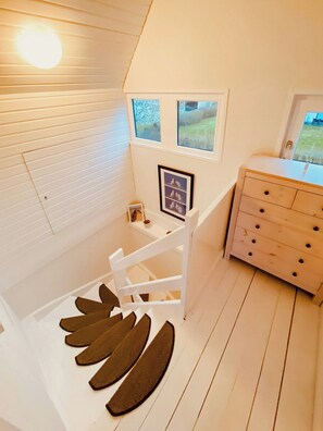 Ein Zimmer mit schrägem Dach, einem Holzkasten, einem gerahmten Bild und einem Fenster mit Blick auf Grünflächen.