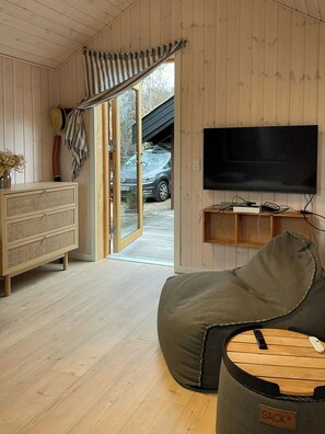 Ein gemütliches Zimmer mit Holzboden, einem an der Wand befestigten Fernseher, einer Couch, einem Korbtisch und einer Tür, die nach draußen führt.
