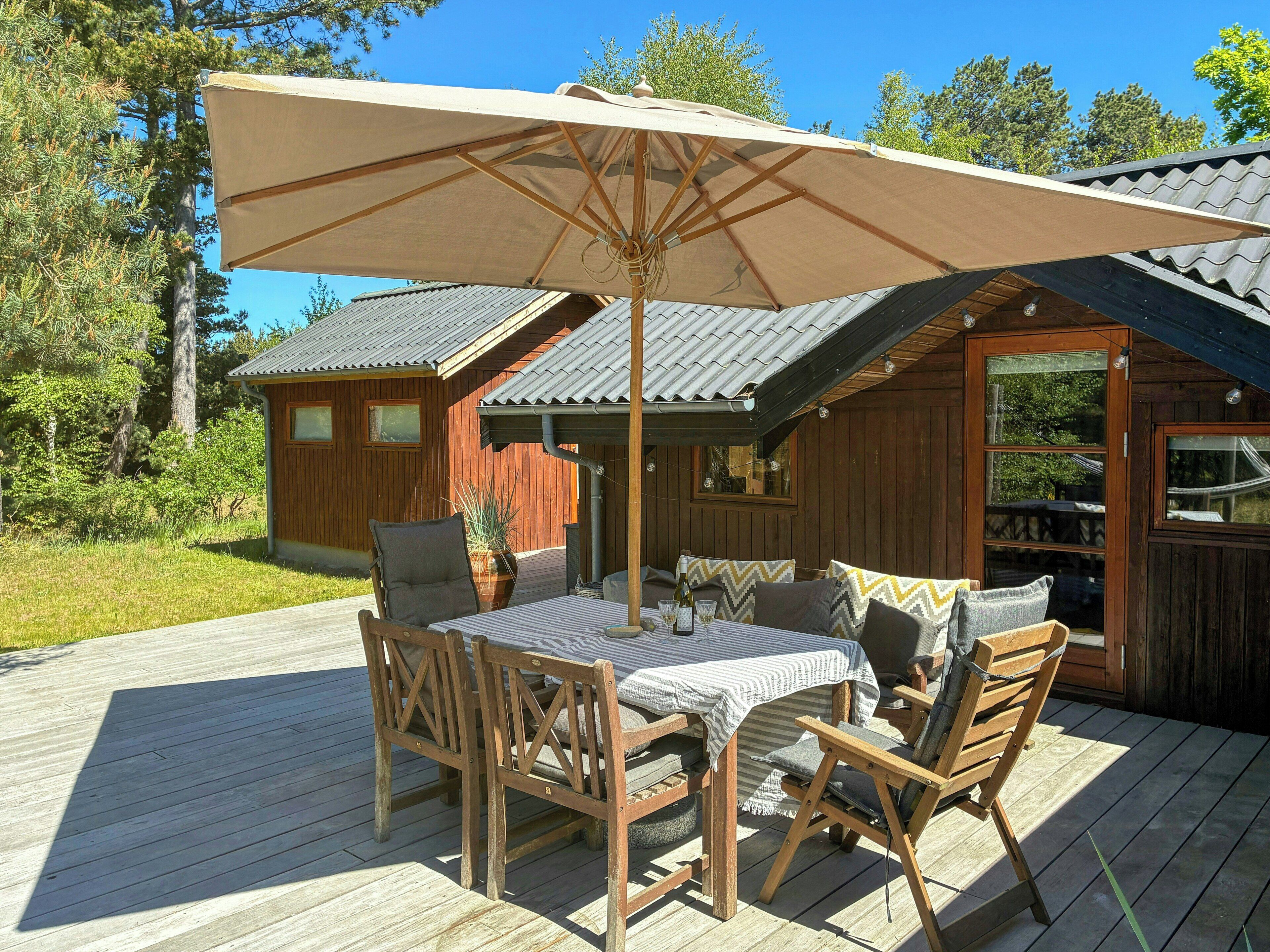 Een houten terras met een tafel, stoelen en een grote parasol, plus een gebouw met houten wanden en ramen.