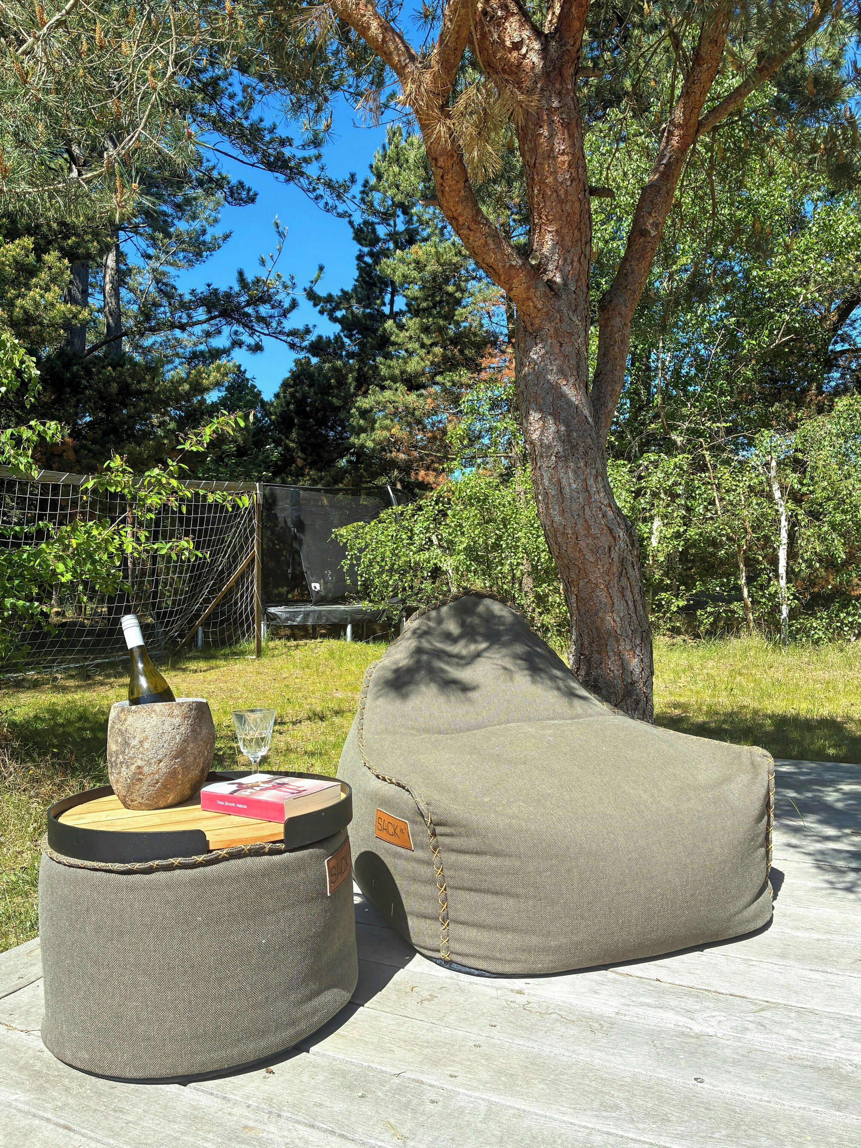 A stone mortar with a bottle and a glass, a bean bag chair, and a wooden table outdoors with trees in the background.