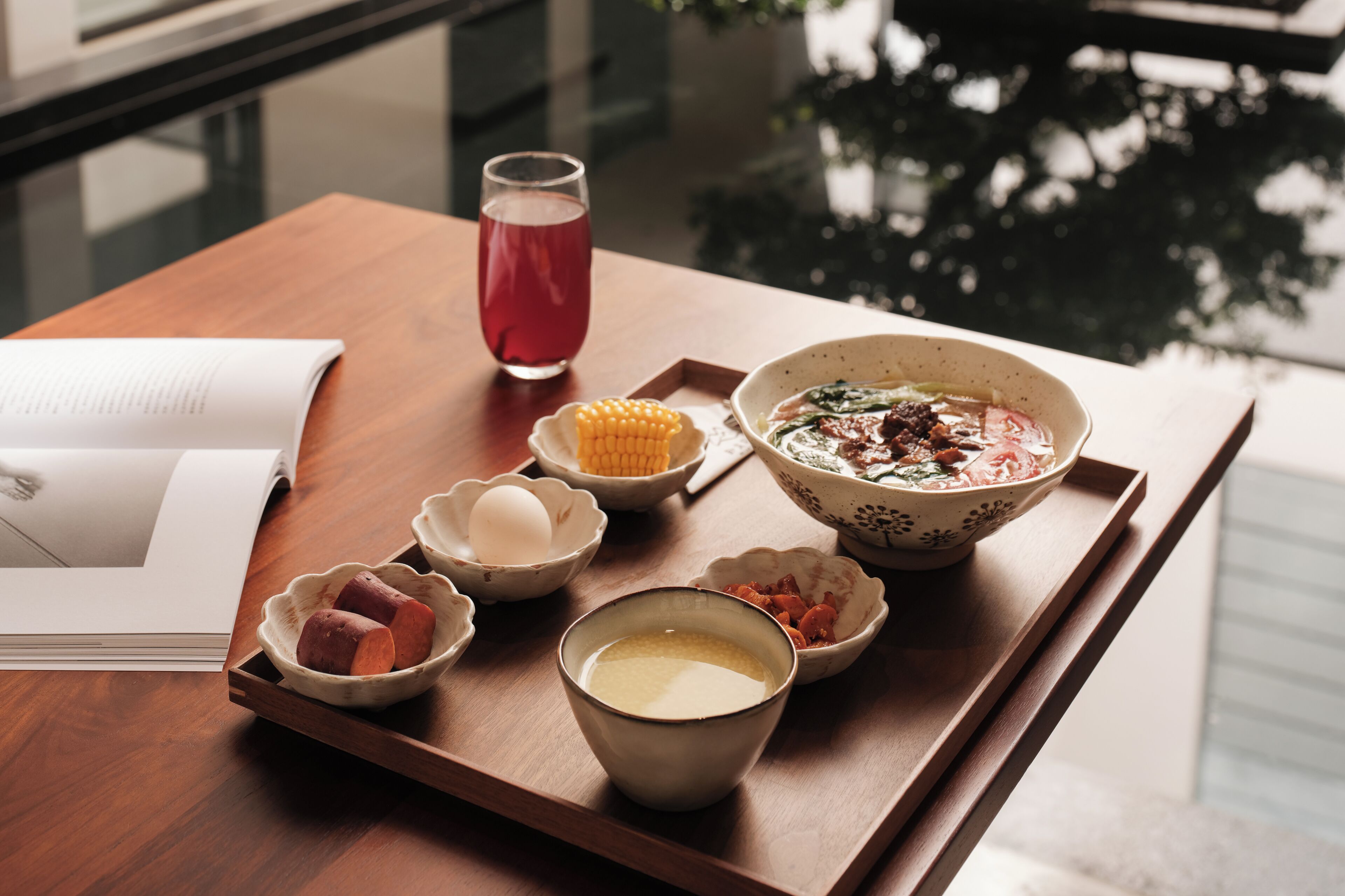 A tray with various bowls of food, a glass of red drink, and a book on a wooden table.
