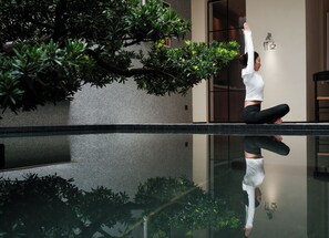 Uma pessoa meditando em um lago espelhado, com um edifício e vegetação ao fundo.