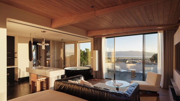 A modern hotel room with a large bed, a sitting area with a sofa and a coffee table, a bathroom with a bathtub, and a balcony with a view of the city.