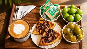 Una bandeja con una taza de café, una porción de tarta, un bol de tomates verdes y una bolsa de galletas.
