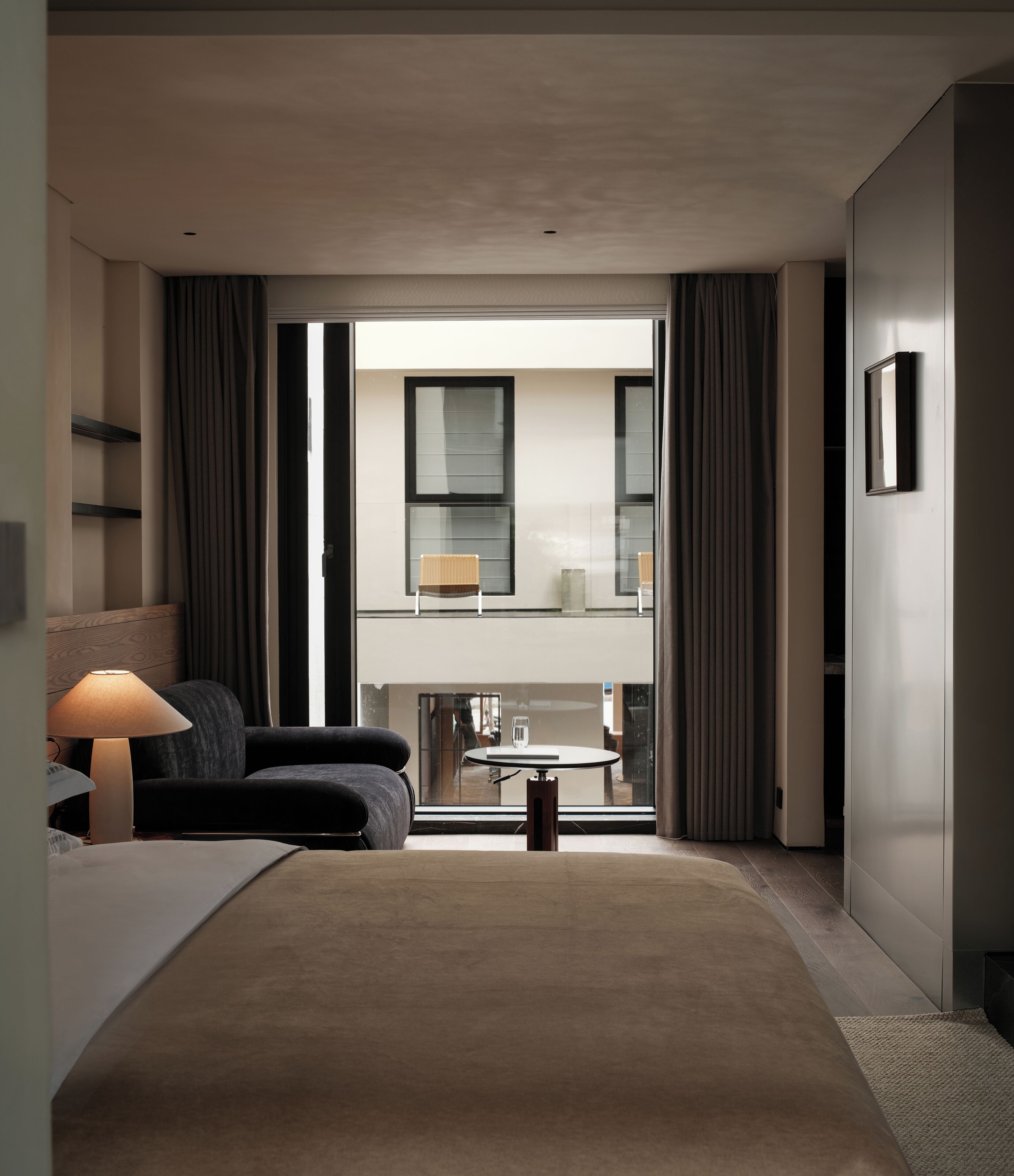 A modern hotel room with a sofa, a small table, a lamp, and a view of the balcony.