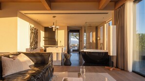 A modern hotel room with a leather sofa, a glass coffee table, a view of a bathroom with a bathtub, and a large window.