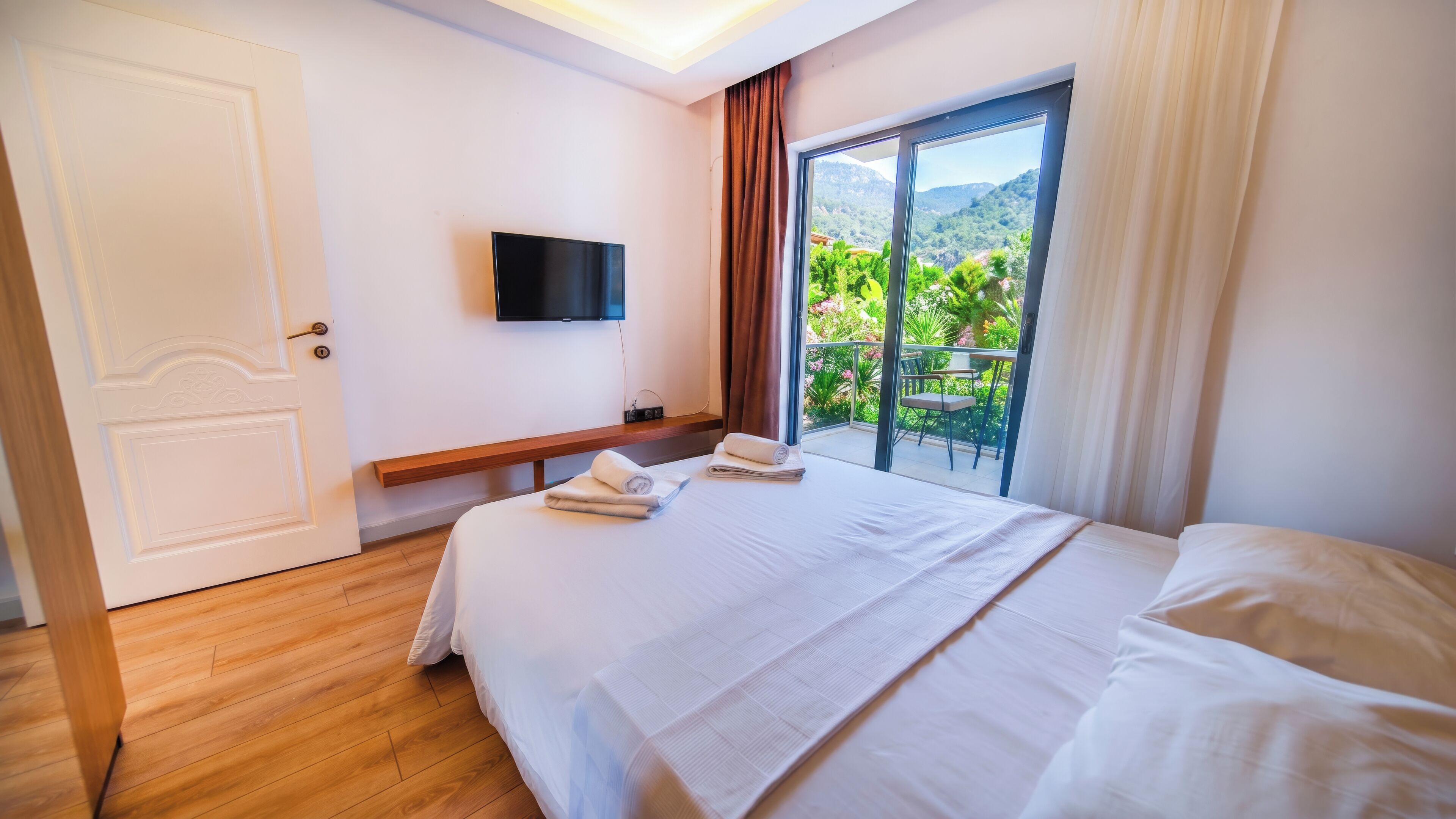 A hotel room with a bed, a television, a balcony with a view of greenery, and a wooden floor.