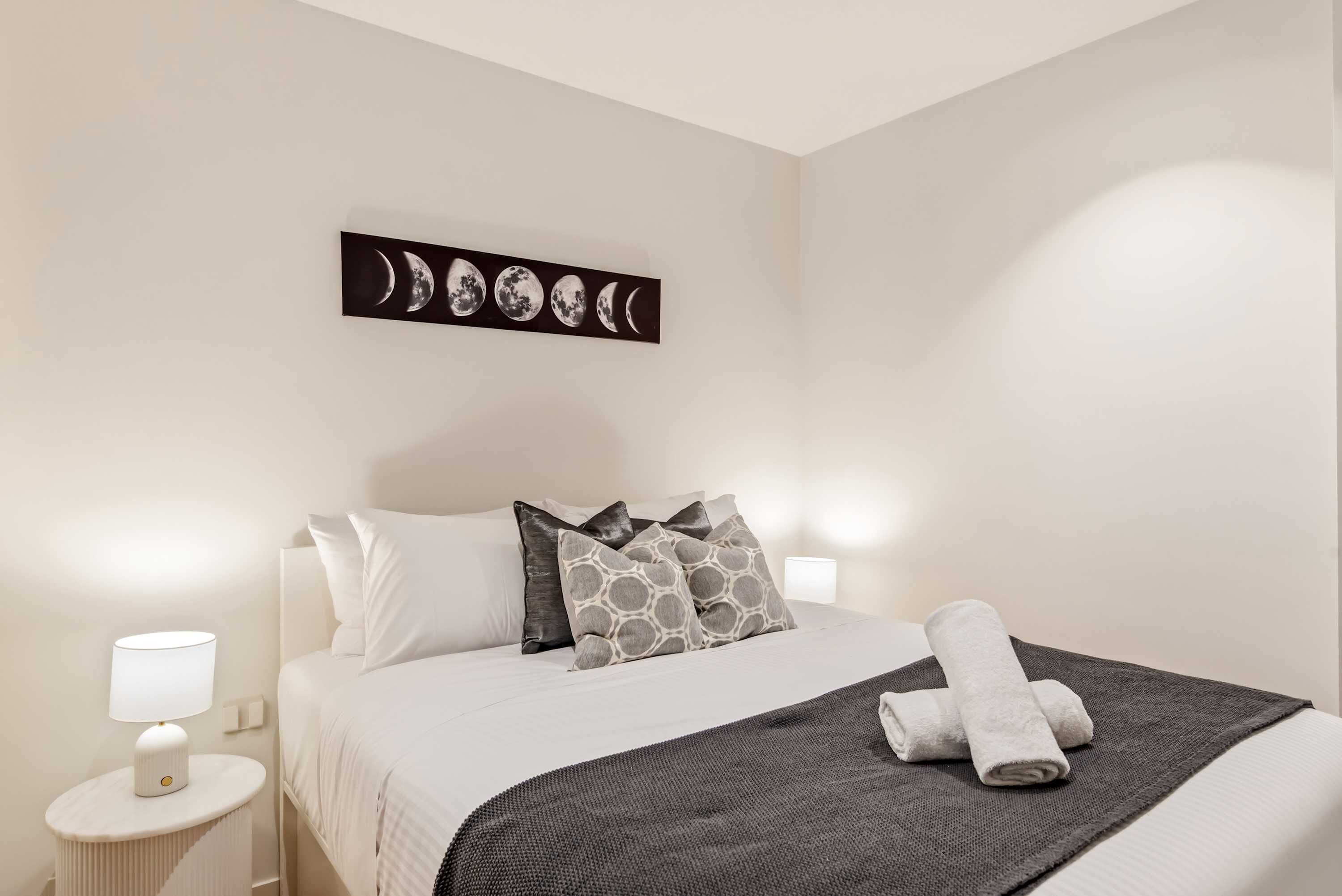 Una cama bien tendida con un edredón estampado, una obra de arte colgada en la pared que representa las fases de la luna y una lámpara de noche.