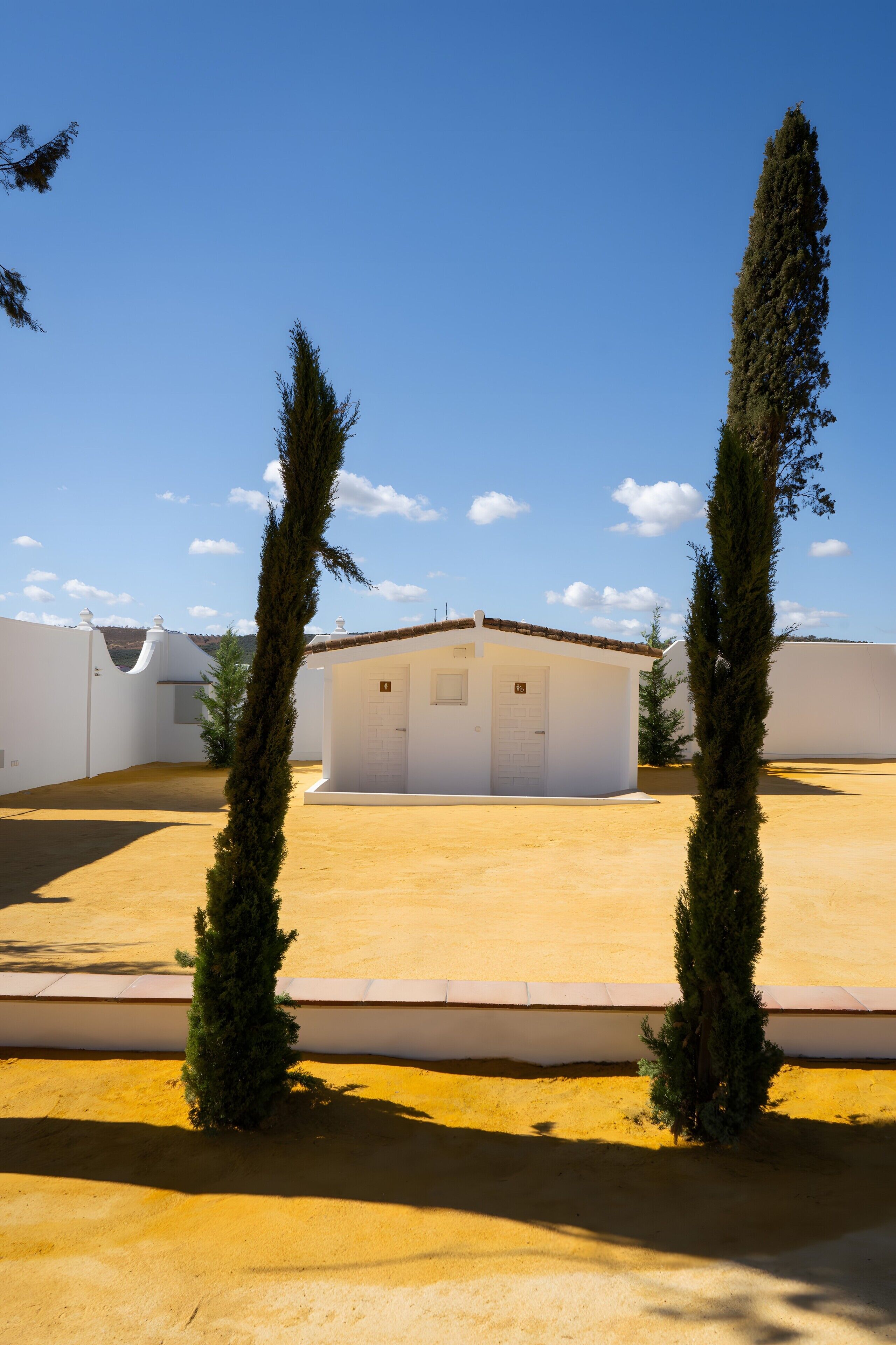 Un edificio blanco con una puerta y ventanas, rodeado de arena y dos árboles altos.