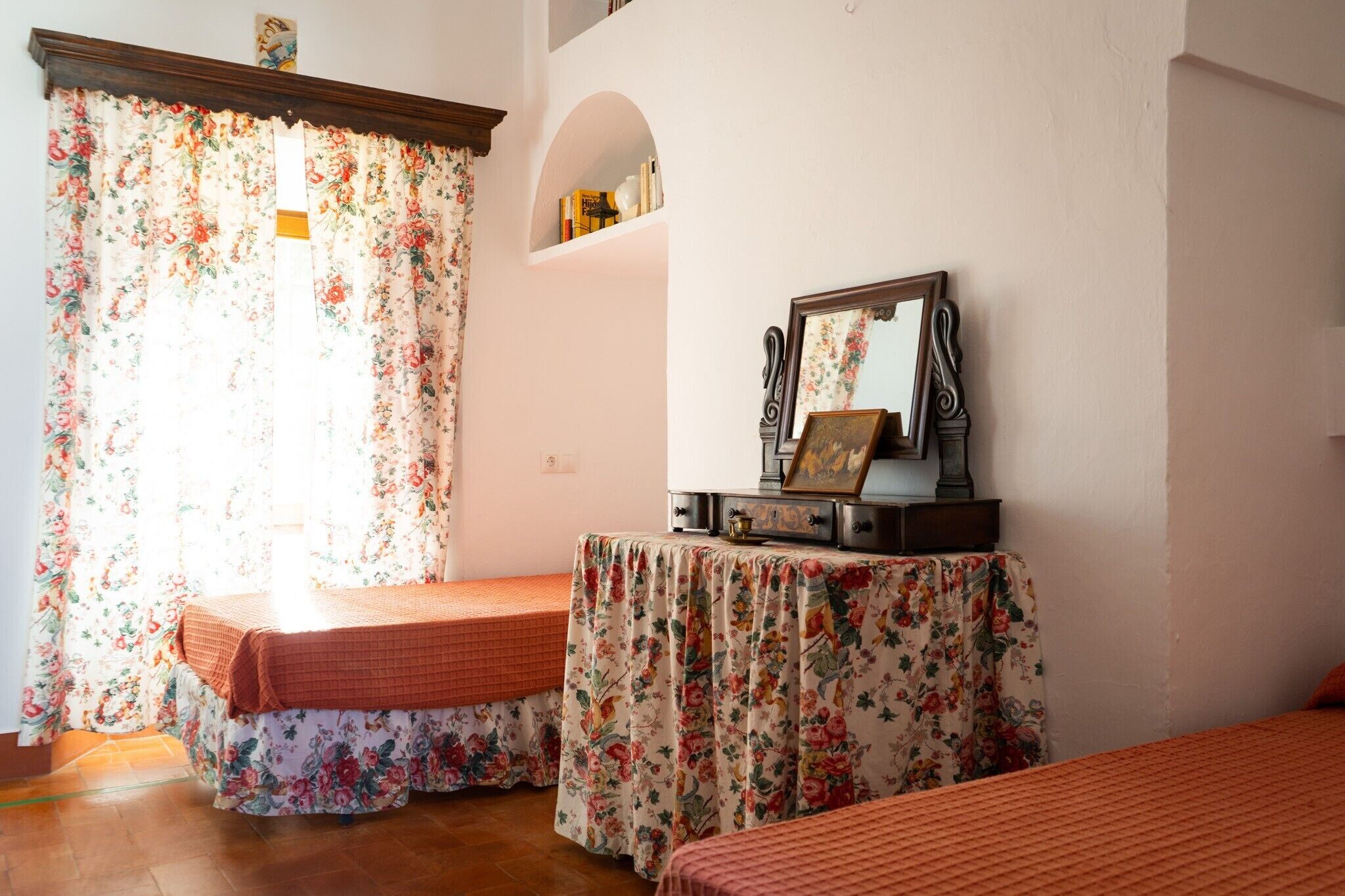 Ein Zimmer mit zwei Betten, blumigen Vorhängen, einem Spiegel und einem kleinen Tisch mit einer Lampe.
