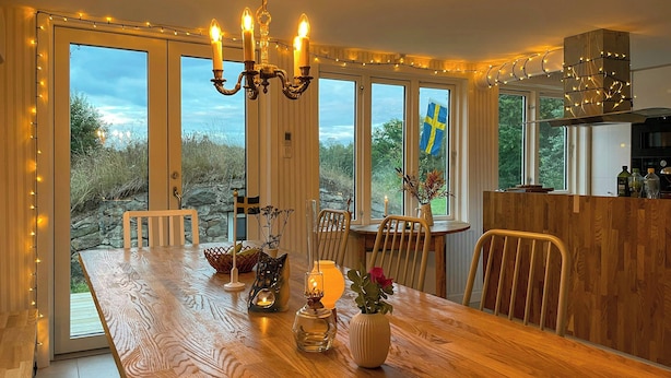 Spiseområde med et træbord, stole, en lysekrone og udsigt til det fri.