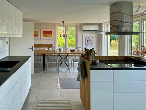 Eine moderne Küche mit weißen Schränken, ein Essbereich mit Holztisch und -stühlen sowie große Fenster mit Blick ins Grüne.