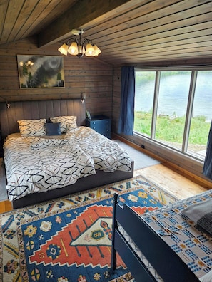 A bedroom with a large bed, a view of a lake, a wooden ceiling, and a painting on the wall.