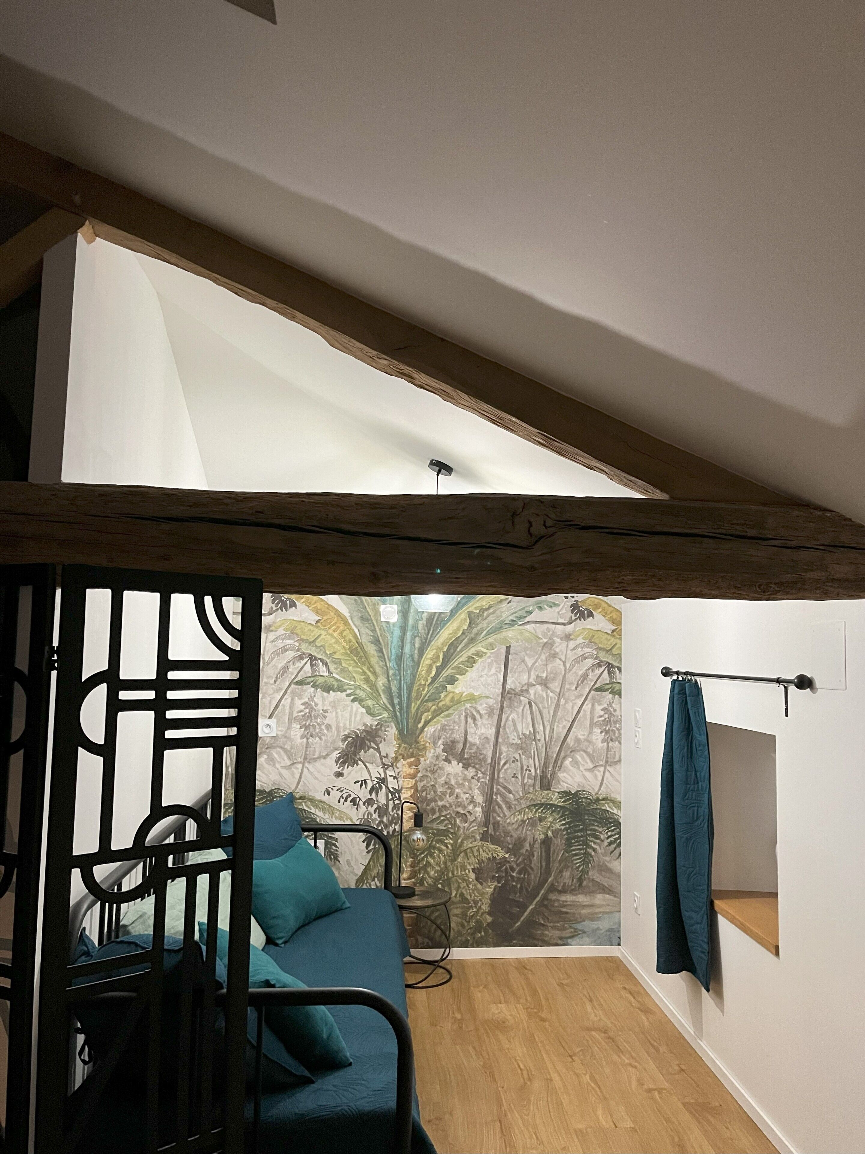 A bedroom with a bed, a window with a view of foliage, a wooden ceiling, and a wall-mounted light fixture.