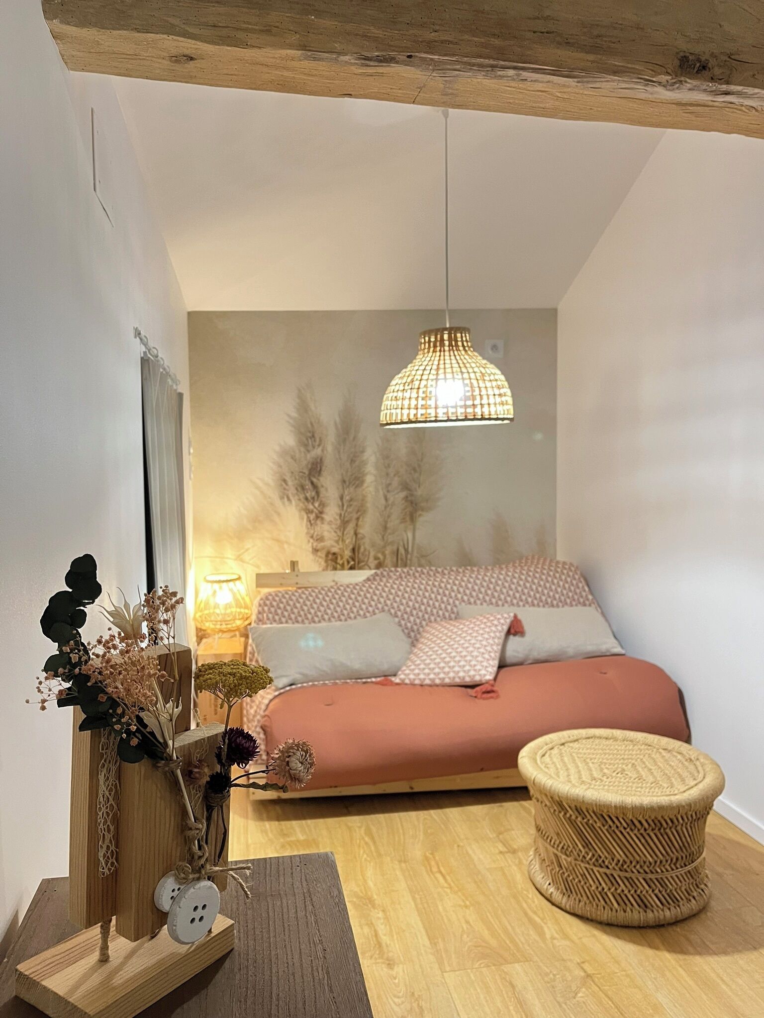 Ein Schlafzimmer mit einem Bett, einer Pendelleuchte, einem Nachttisch und einem gewebten Hocker.