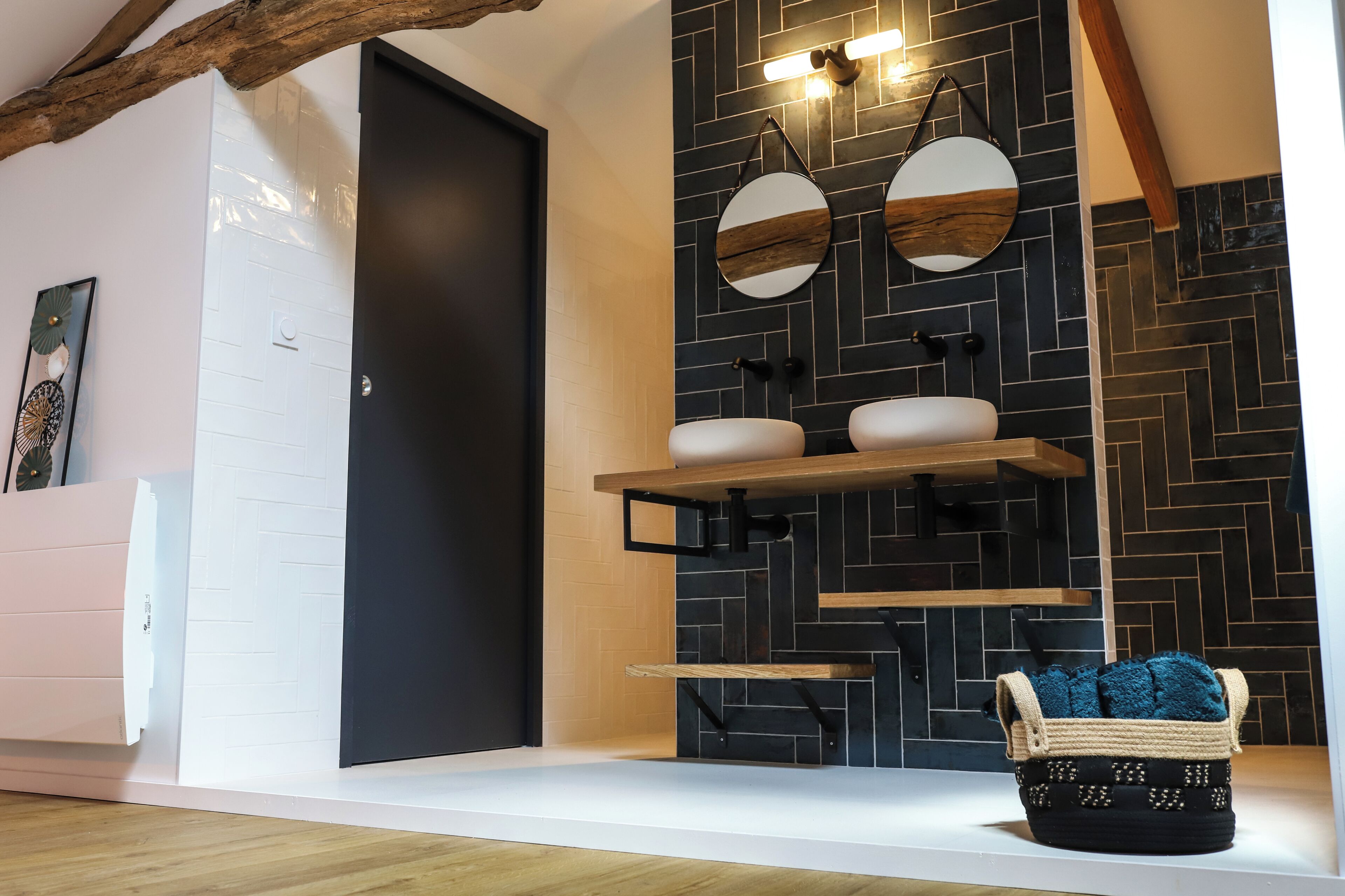 A modern bathroom with a black door, white walls, and a herringbone-patterned black tile backsplash. There are two round mirrors with wooden frames, a white sink, and a wooden shelf above the sink.