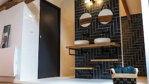 Ein modernes Badezimmer mit einer schwarzen Tür, weißen Wänden und einer schwarzer Fliesenwand im Fischgrätmuster. Zwei runde Spiegel mit Holzrahmen, ein weißes Waschbecken und ein Holzregal darüber.
