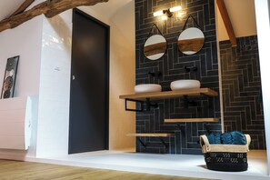 Ein modernes Badezimmer mit einer schwarzen Tür, weißen Wänden und einer schwarzer Fliesenwand im Fischgrätmuster. Zwei runde Spiegel mit Holzrahmen, ein weißes Waschbecken und ein Holzregal darüber.