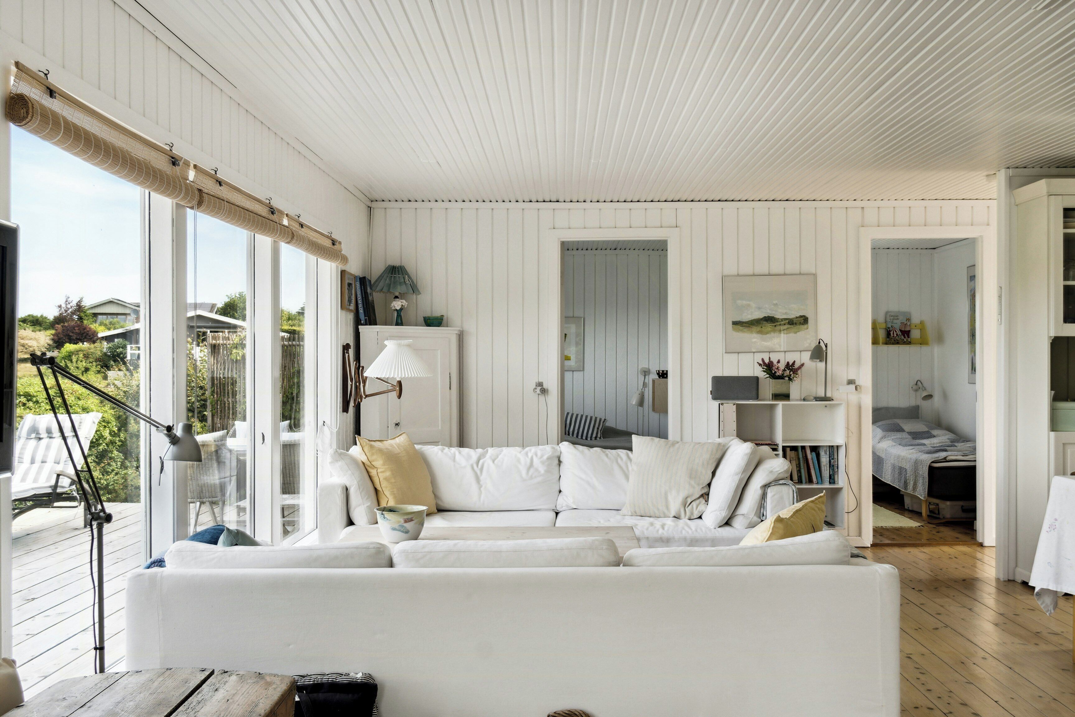 Un intérieur blanc avec une grande fenêtre, un canapé blanc et une terrasse en bois visible à l’extérieur.