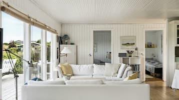 Un intérieur blanc avec une grande fenêtre, un canapé blanc et une terrasse en bois visible à l’extérieur.
