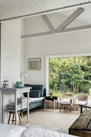 A room with a bed, a desk, a chair, a sofa, and a large window with a view of greenery. - Sunset over Sejerø Bay from house (Fårevejle)