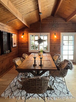 A wooden interior with a dining table, chairs, pendant lights, and a window with a view. - Authentic log cabin on large enclosed natural plot (Vig)