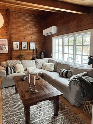 A cozy cabin interior with a wooden ceiling, a large window, a sofa set, and a table with candles. - Authentic log cabin on large enclosed natural plot (Vig)