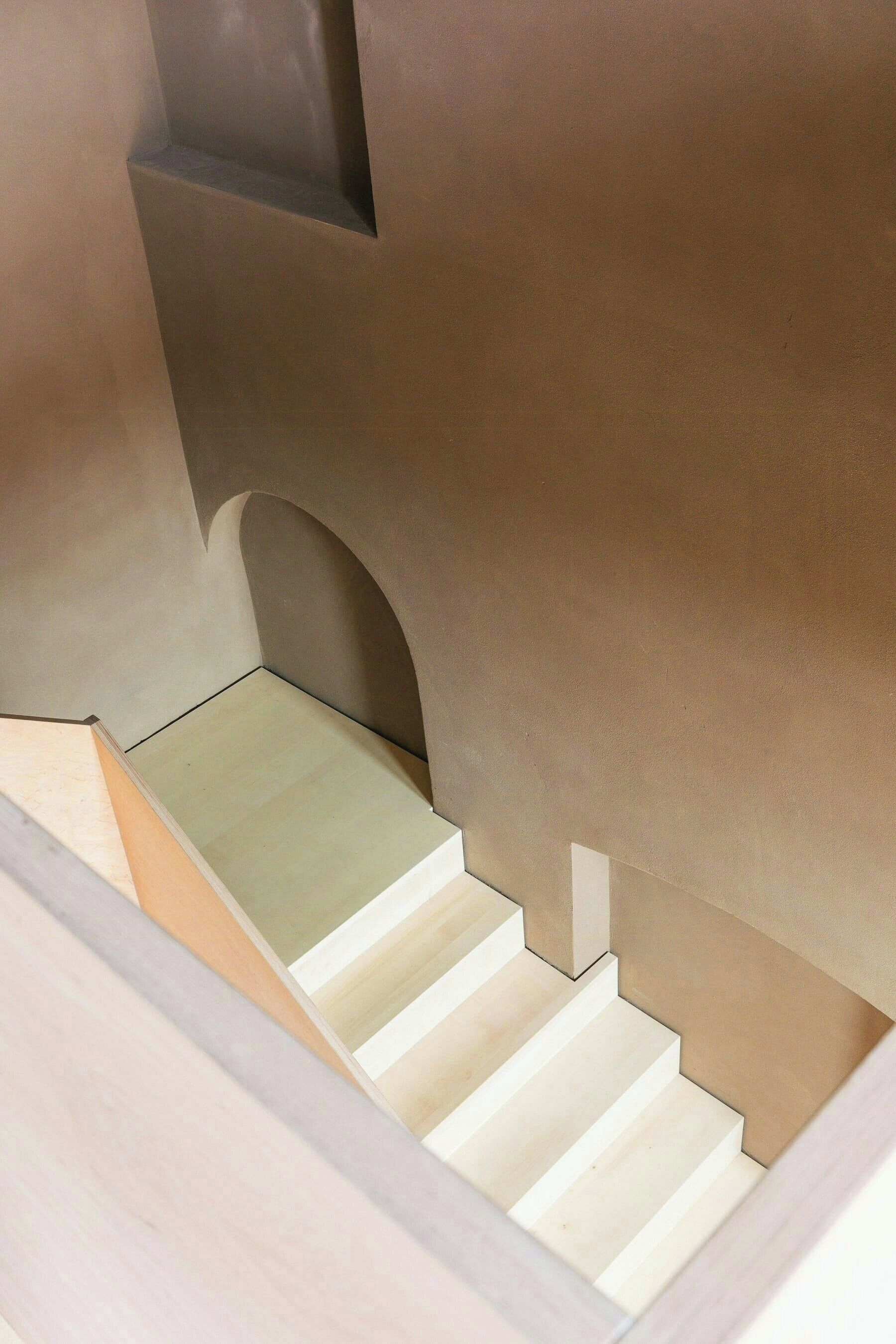 Eine Treppe mit hellfarbener Handlauf und Stufen, die zu einer Zwischenebene mit dunkler Wand und einer kleinen Nische führt.