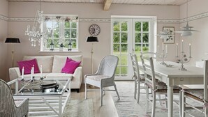 A room with a white sofa, a glass coffee table, a wicker chair, a dining table with chairs, and a chandelier. - Guest house by lake and forest (Nakskov)
