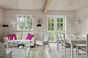A room with a white sofa, a glass coffee table, a wicker chair, a dining table with chairs, and a chandelier. - Guest house by lake and forest (Nakskov)
