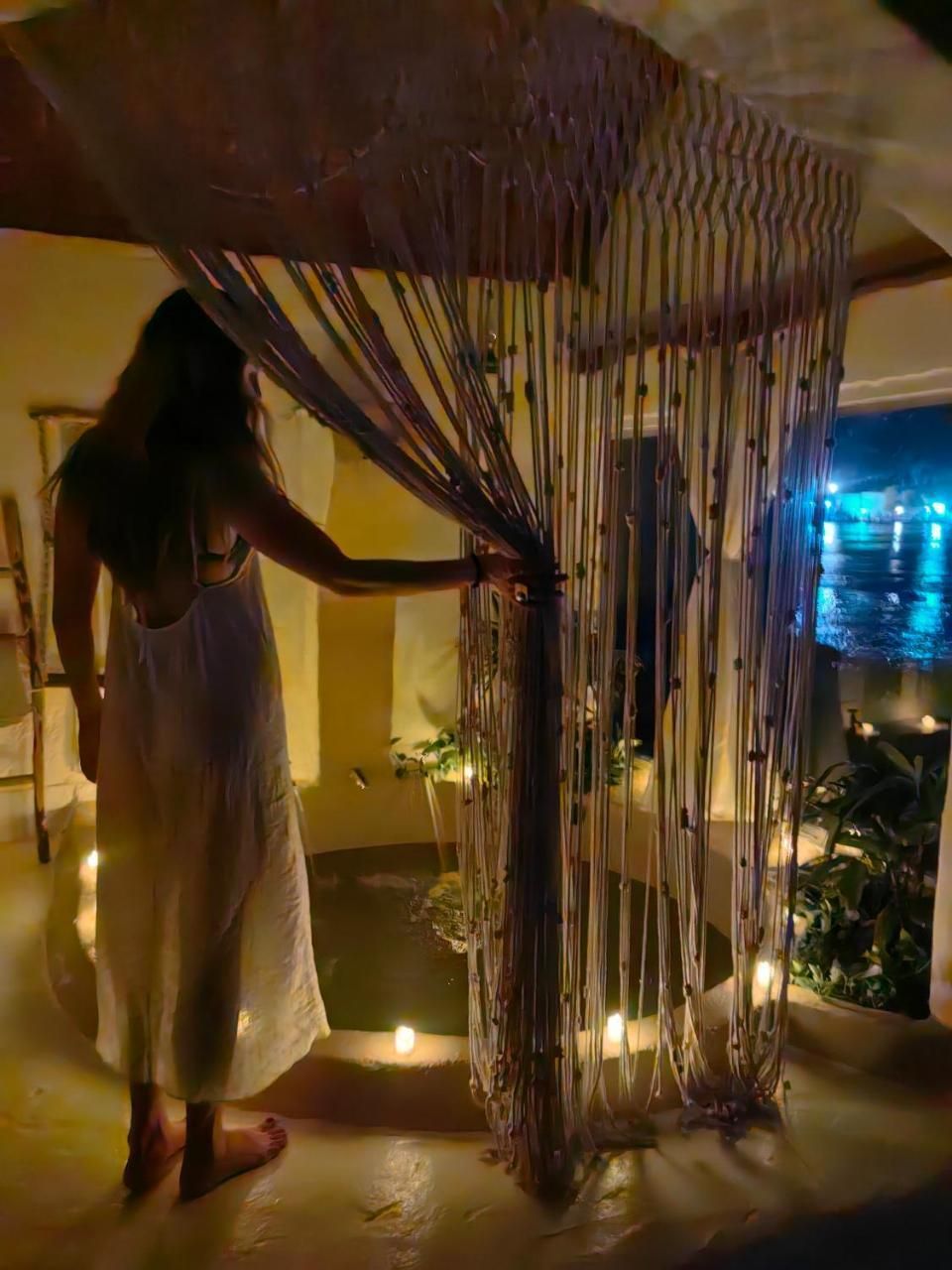 A person in a white dress standing under a canopy with vertical strings, in a room with a view of a body of water.