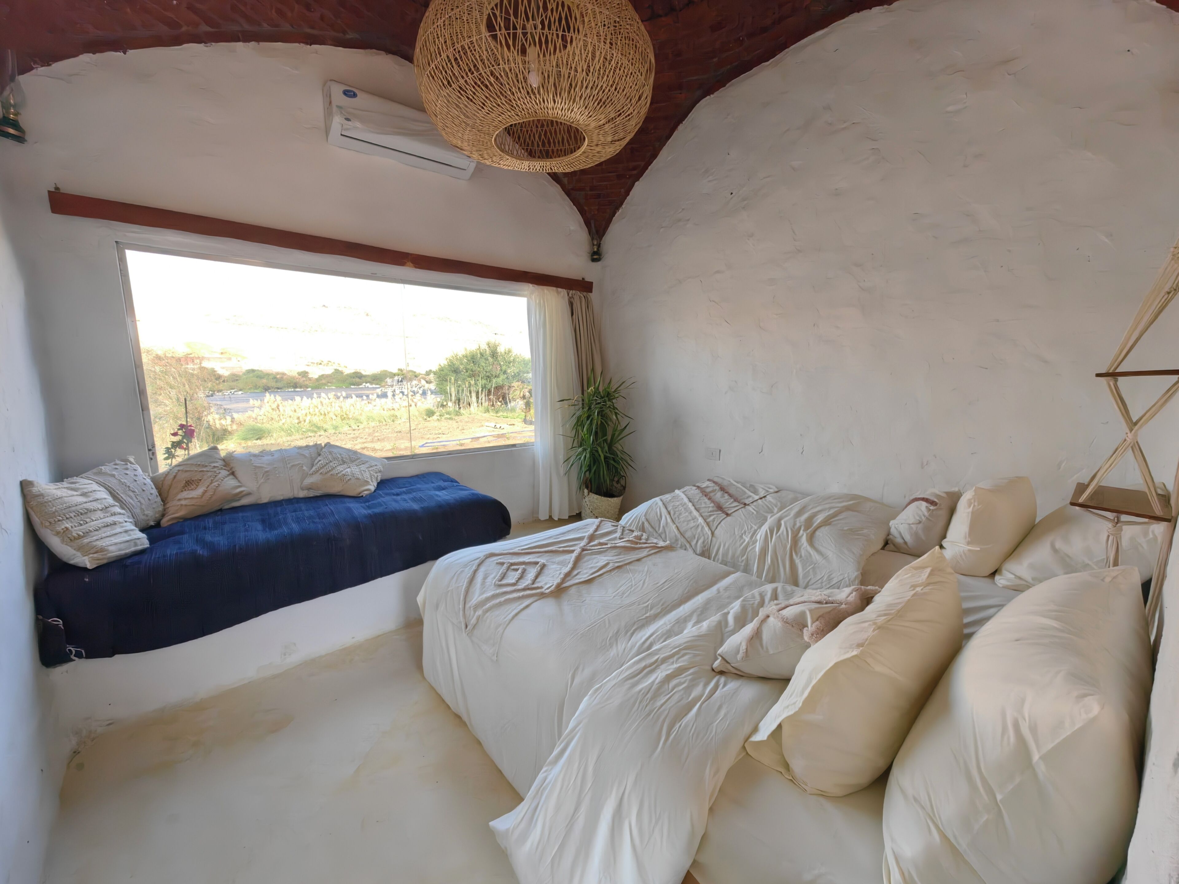 Une chambre avec une grande fenêtre, un lit avec de nombreux oreillers, un luminaire suspendu et un climatiseur fixé au mur.