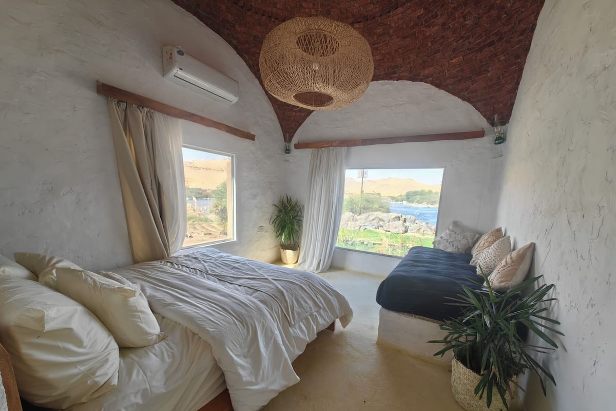 Une chambre avec un grand lit, un plus petit lit, une plante en pot, un luminaire suspendu et une vue sur un paysage.